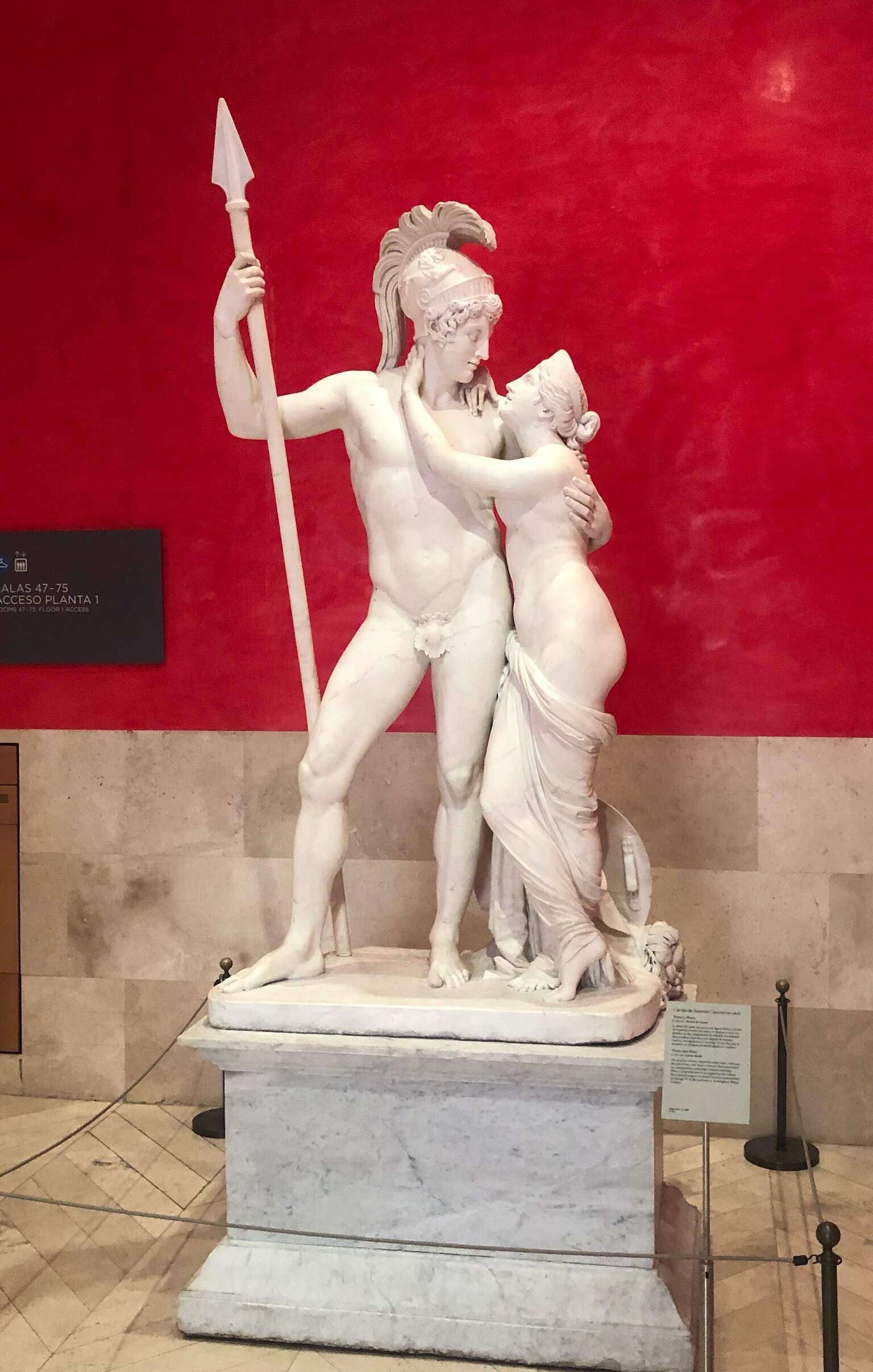 White Statue in the Prado Museum in Spain with bright red walls showing a man holding a spear and a woman with her arms around his neck